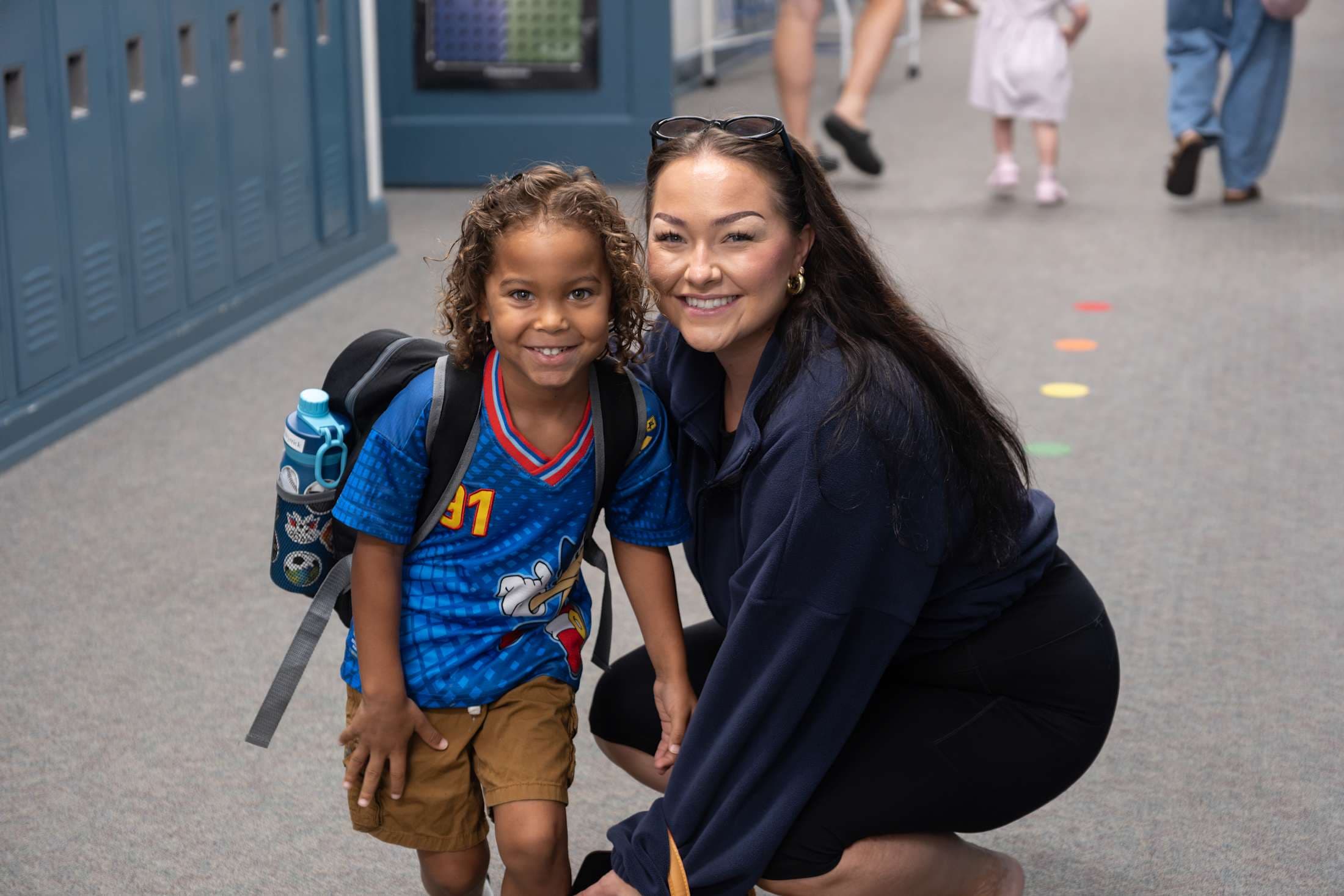 A smiling woman kneels beside a young boy with a backpack in a school hallway with lockers and other children in the background.