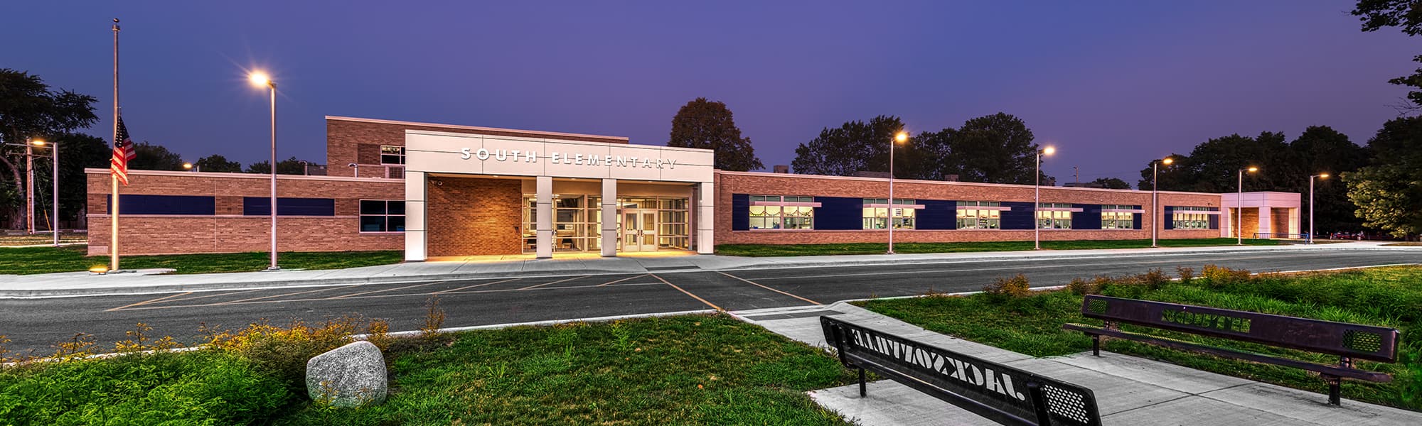 A wide view of South Elementary shows the modern brick school building at dusk, with a brightly lit main entrance, landscaped grounds, an American flag, and benches in the foreground overlooking the driveway.