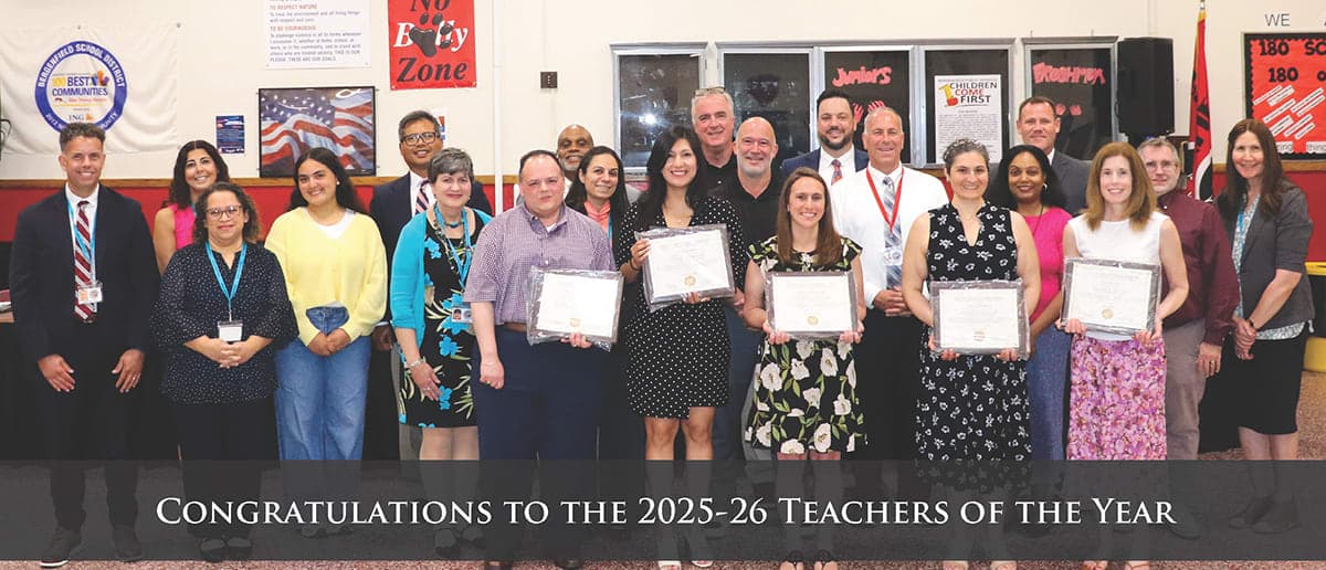 A group of teachers stand in the foreground holding certificates, with a school cafeteria decorated with banners and posters in the background.