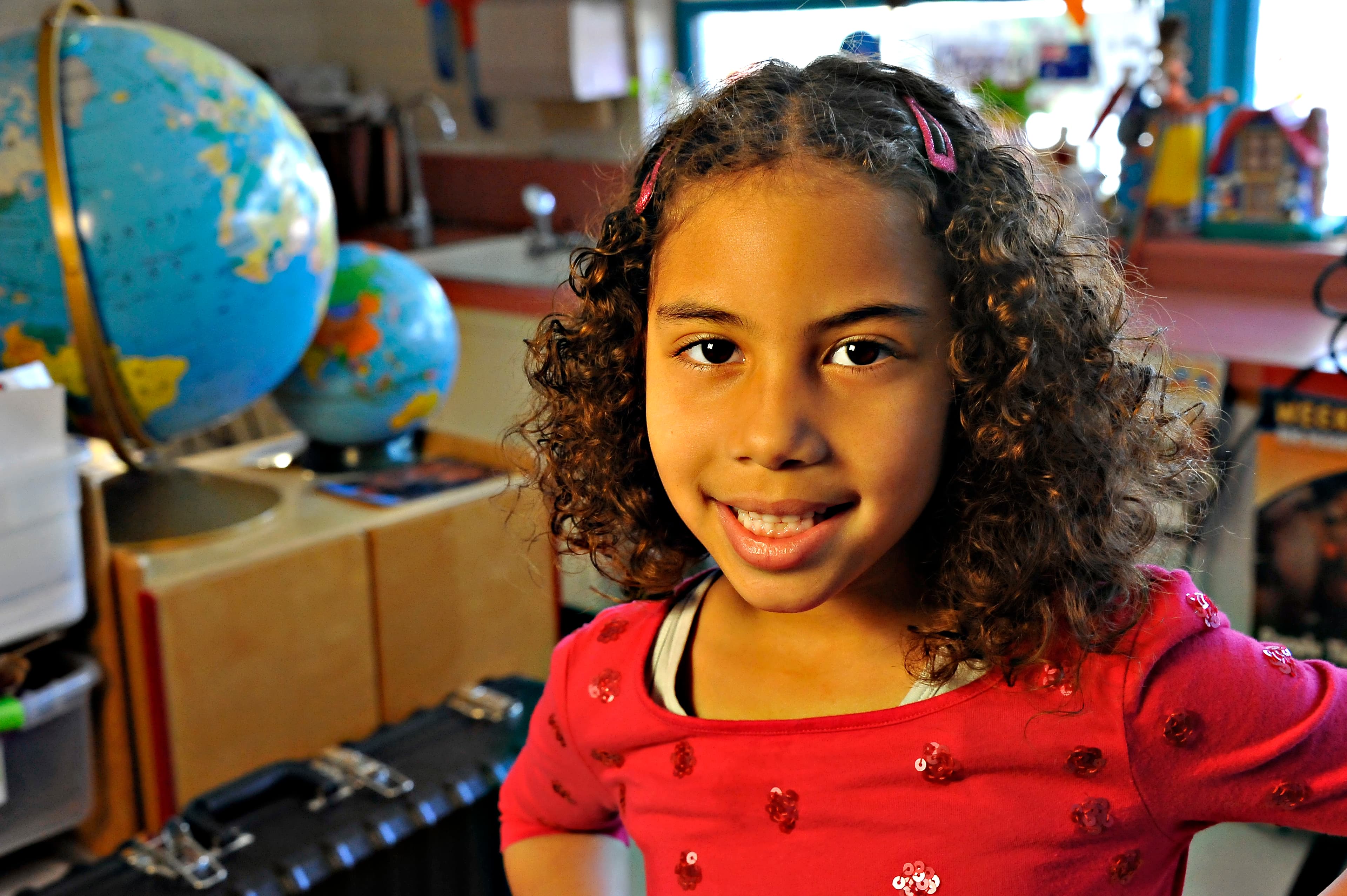 A little girl in a red shirt smiles in front of two globes in the classroom