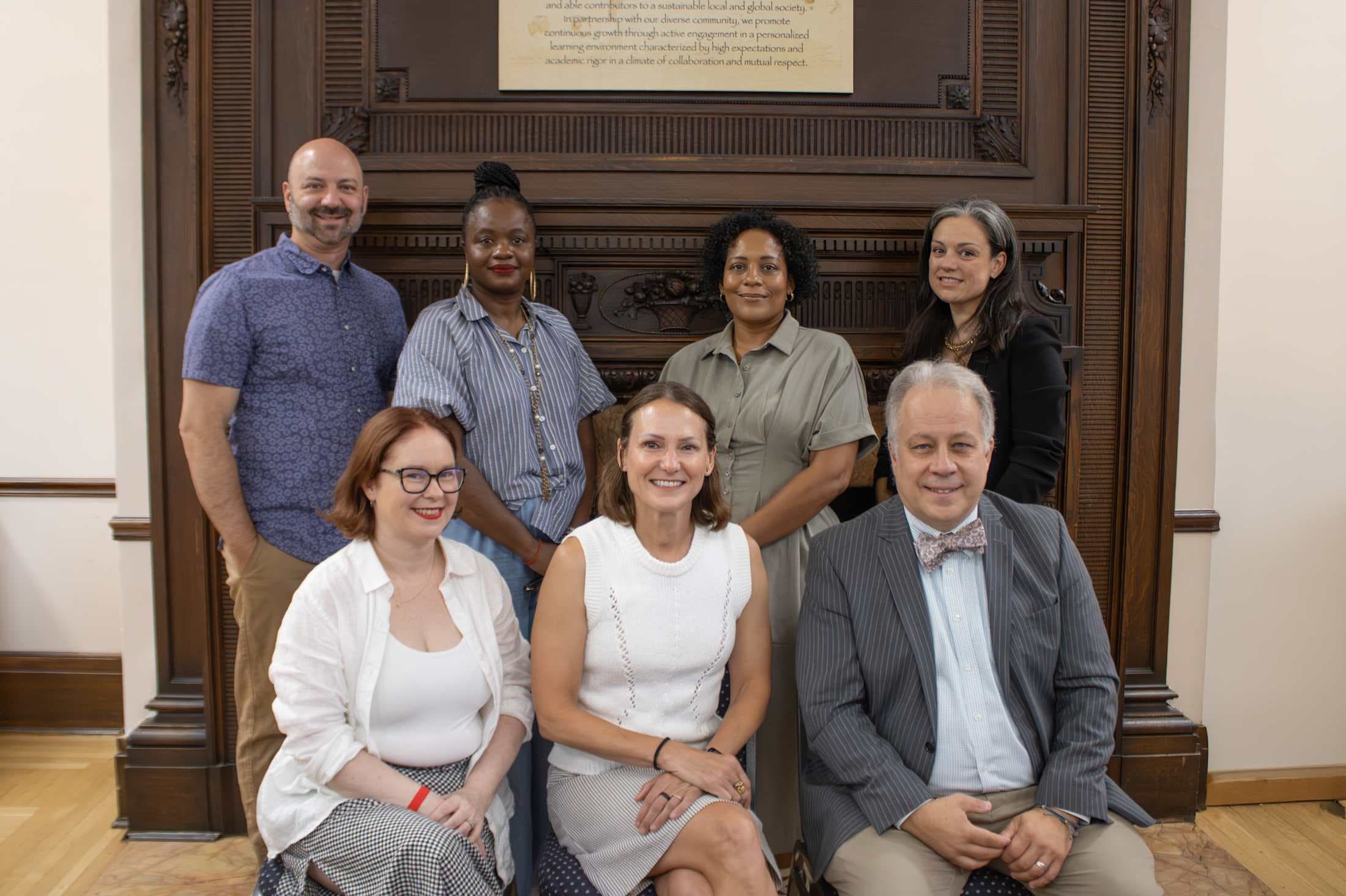 Seven people pose for a photo in front of a large, ornate fireplace with a framed document above it.