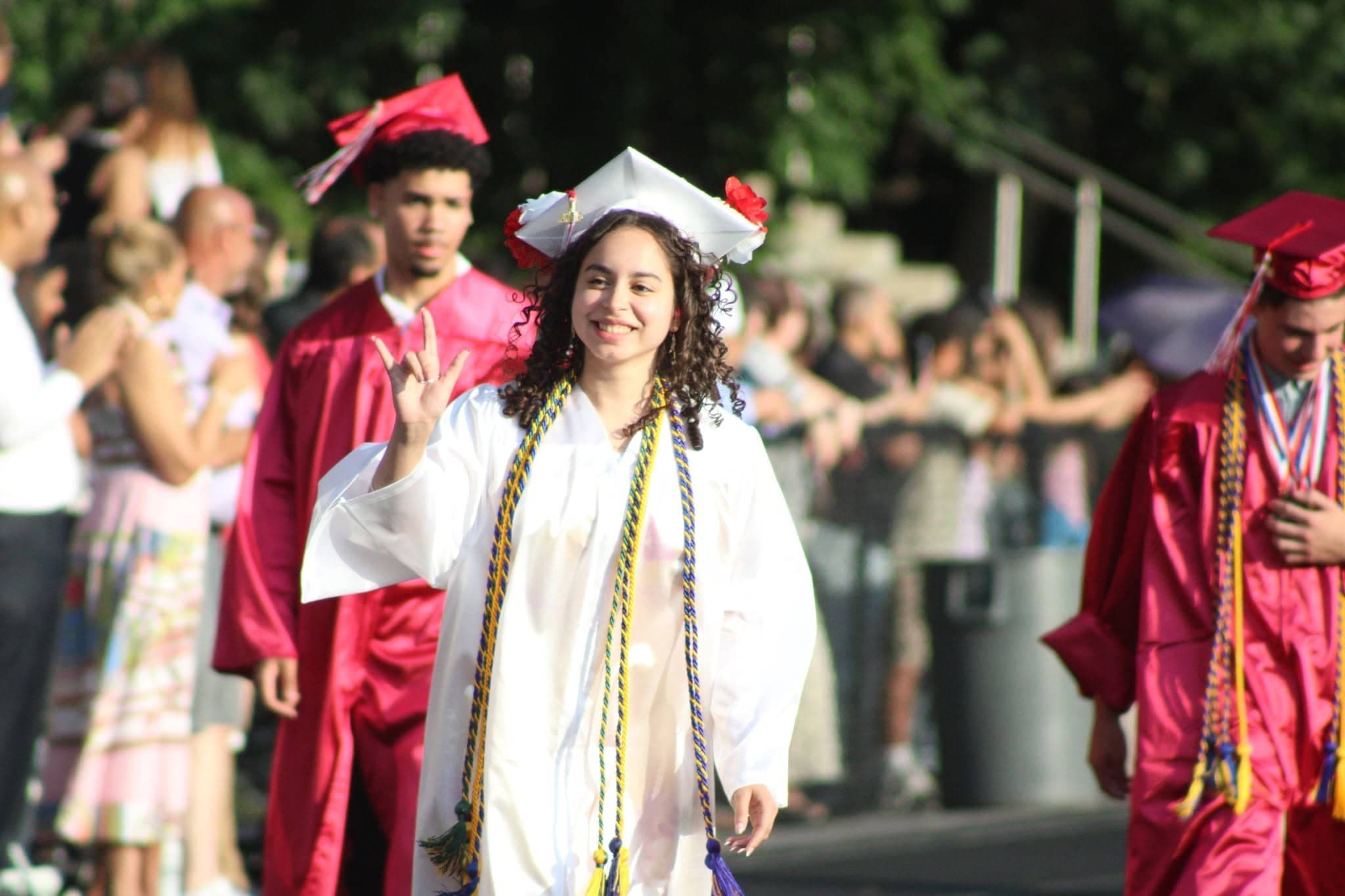 A smiling graduate in a white cap and gown walks forward in the foreground, while other graduates in red gowns and blurred figures are visible in the background.