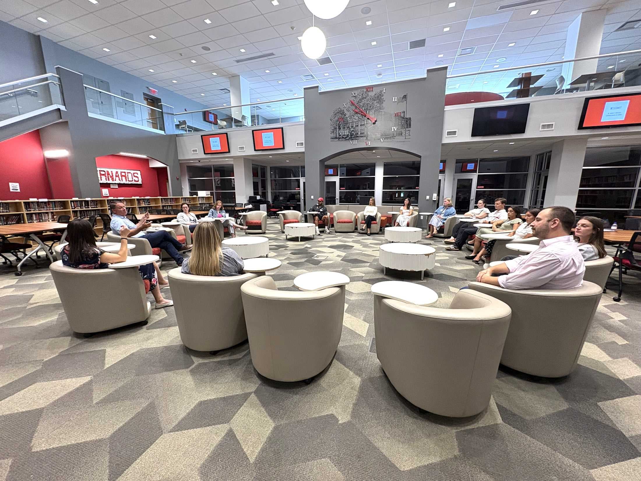 A group of people are seated in a circle of modern chairs in the foreground, while the background reveals a library with bookshelves, a mezzanine, and large screens.