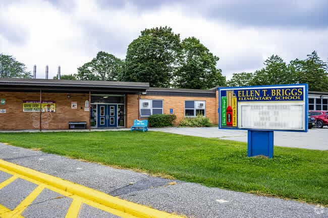 The image shows a brick elementary school building with a sign that reads %22Helen T. Briggs Elementary School%22 surrounded by trees and a grassy lawn.