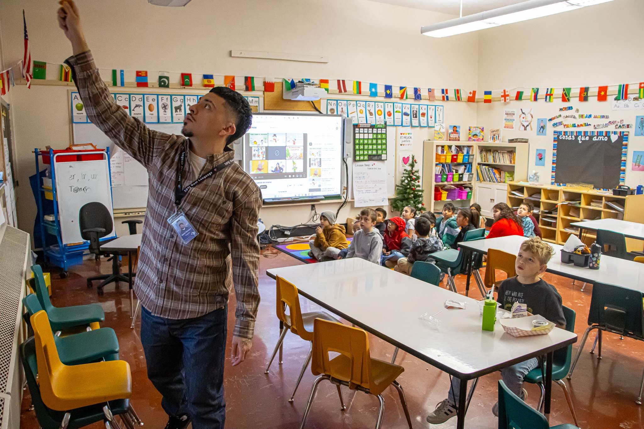 A teacher in a plaid shirt stands in a classroom, reaching up to write on a whiteboard. Students are seated at desks and on the floor in the background, facing a large interactive screen displaying a virtual meeting. The room is brightly decorated with flags from various countries hanging above the chalkboard.