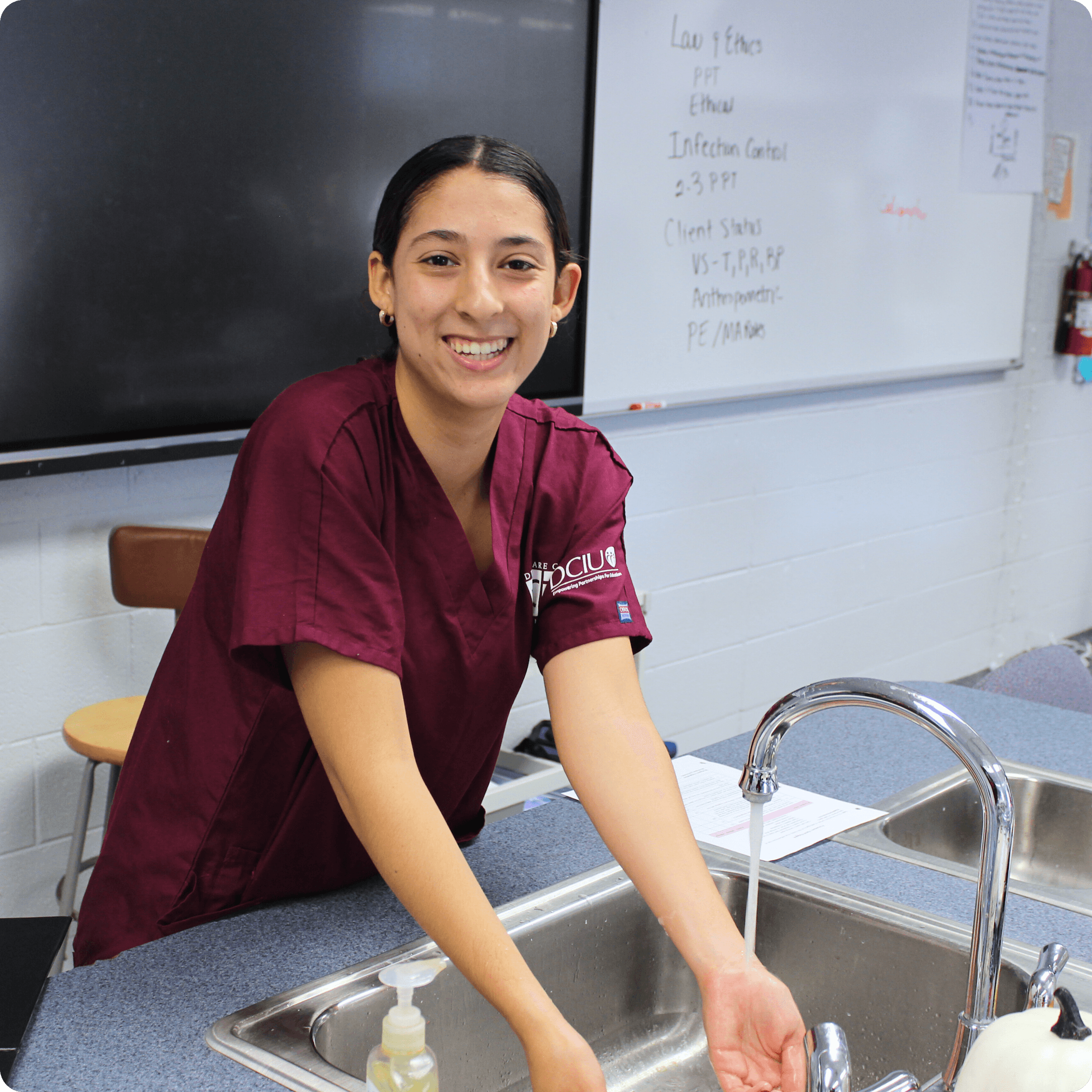 Student in scrubs smiles while washing hands at a sink in the health sciences lab.