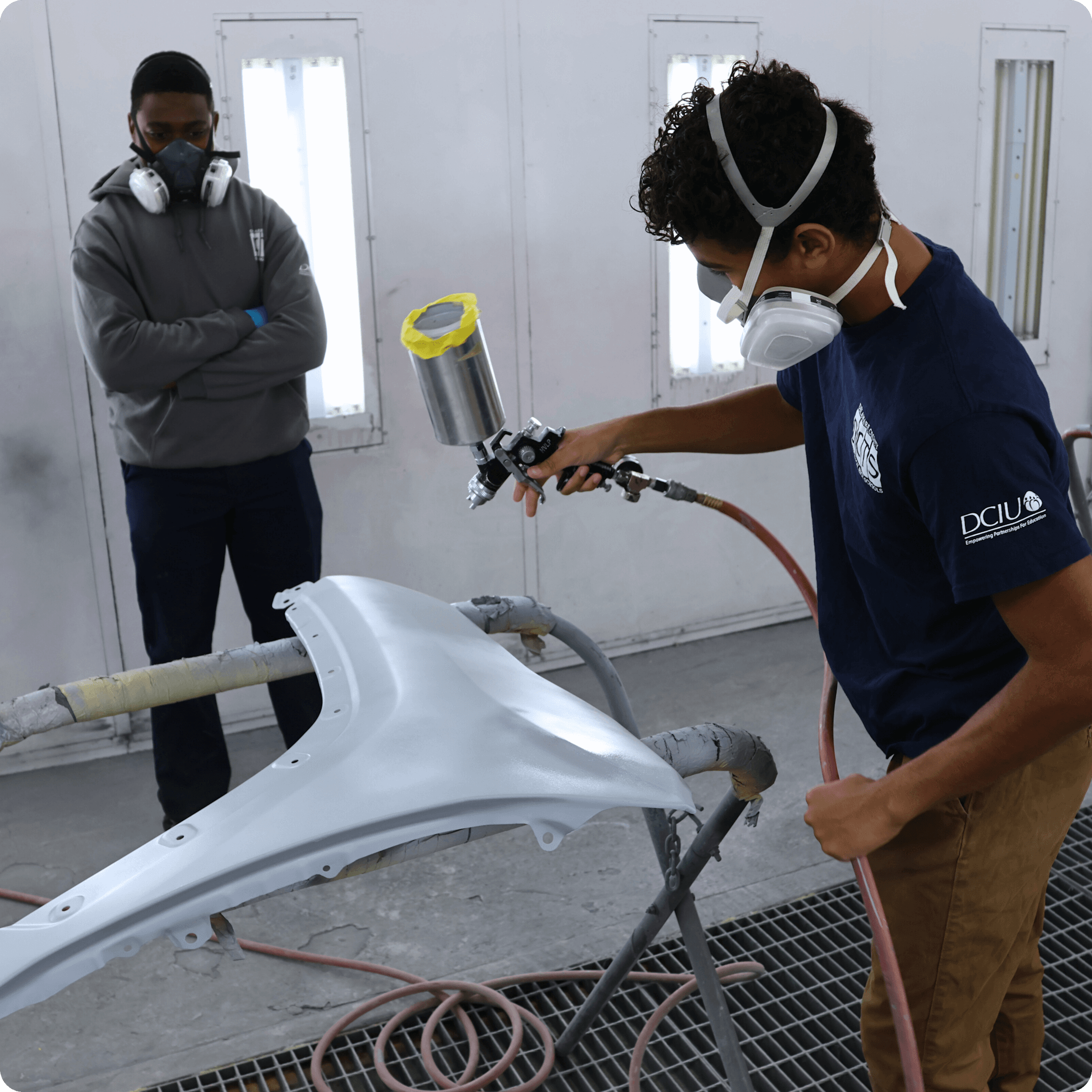 Student sprays paint onto a car part in the collision repair lab while another student observes.