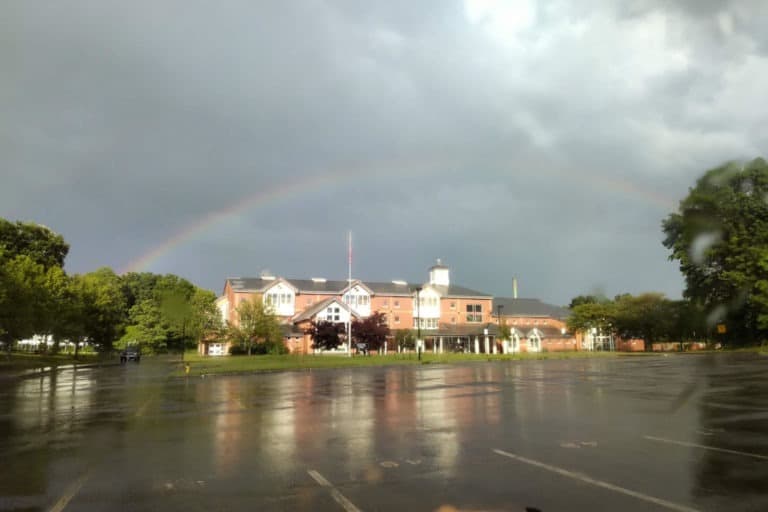 A rainbow arches over a building and a wet parking lot after a rain shower.