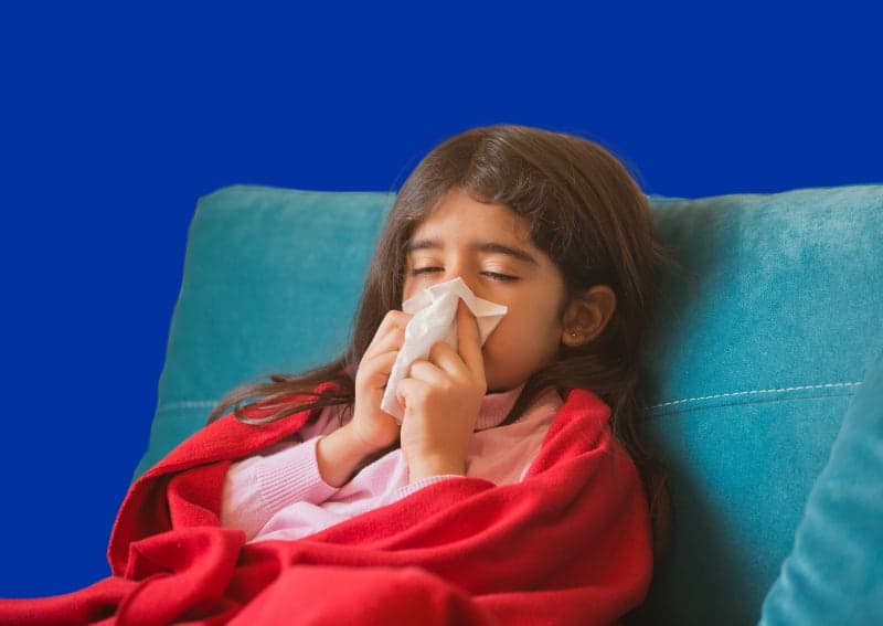 Child ill blowing her nose while sitting on a chair wrapped in a blanket