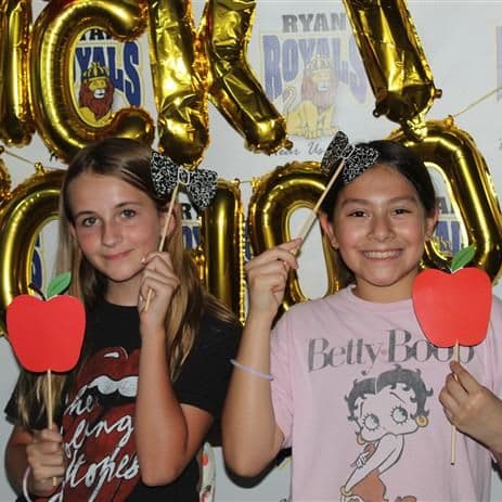 Two young women holding red heart-shaped cutouts in front of their faces, posing in front of a backdrop of large gold balloon letters spelling "ROYALS".