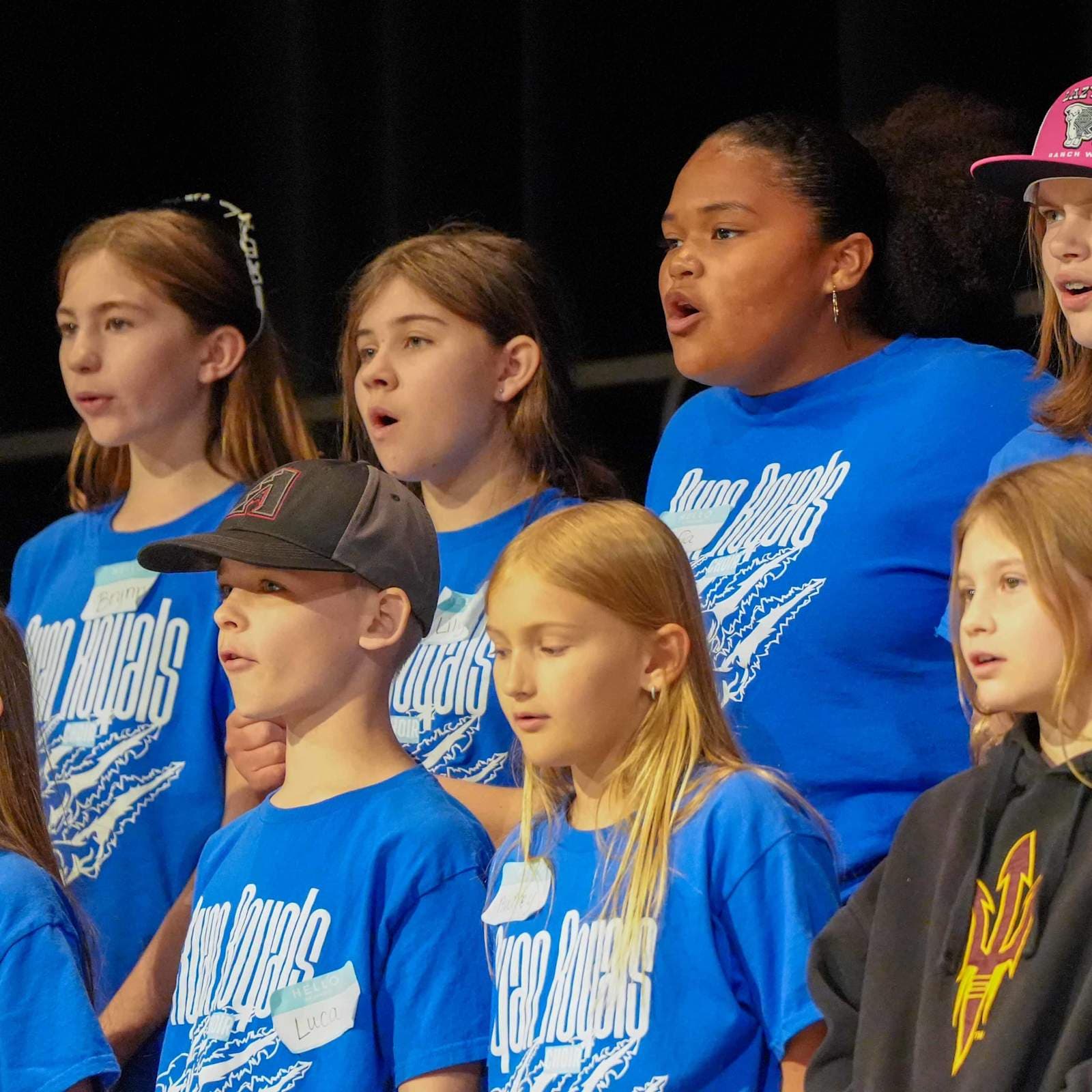 A group of children in blue shirts with "Ocean Angels" written on them sing in front of a dark background.