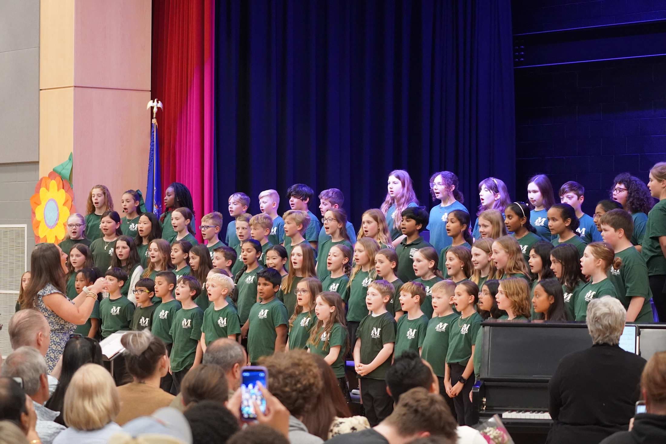 A choir of children in green shirts sings on a stage with a blue curtain backdrop, while an audience watches from the foreground.