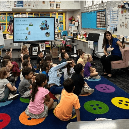 students in a classroom sitting on a colorful rug and teacher reading a book