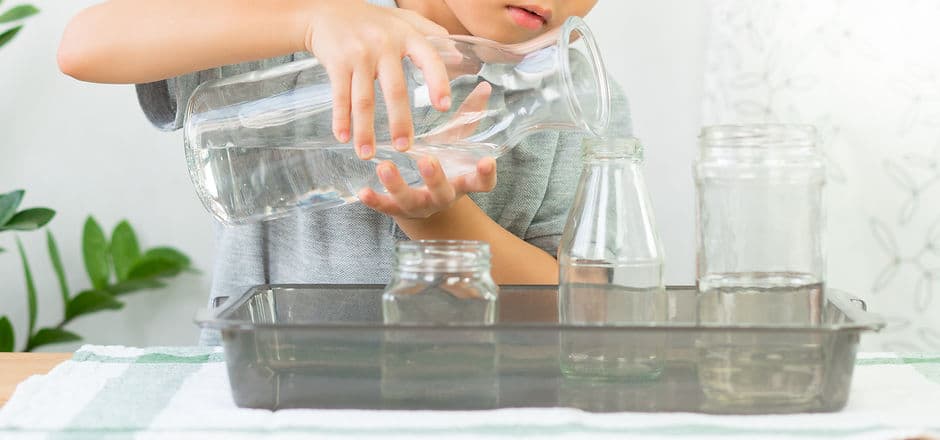 Adorable little kindergarten child, boy pours water from glass jar into bottle with care,
