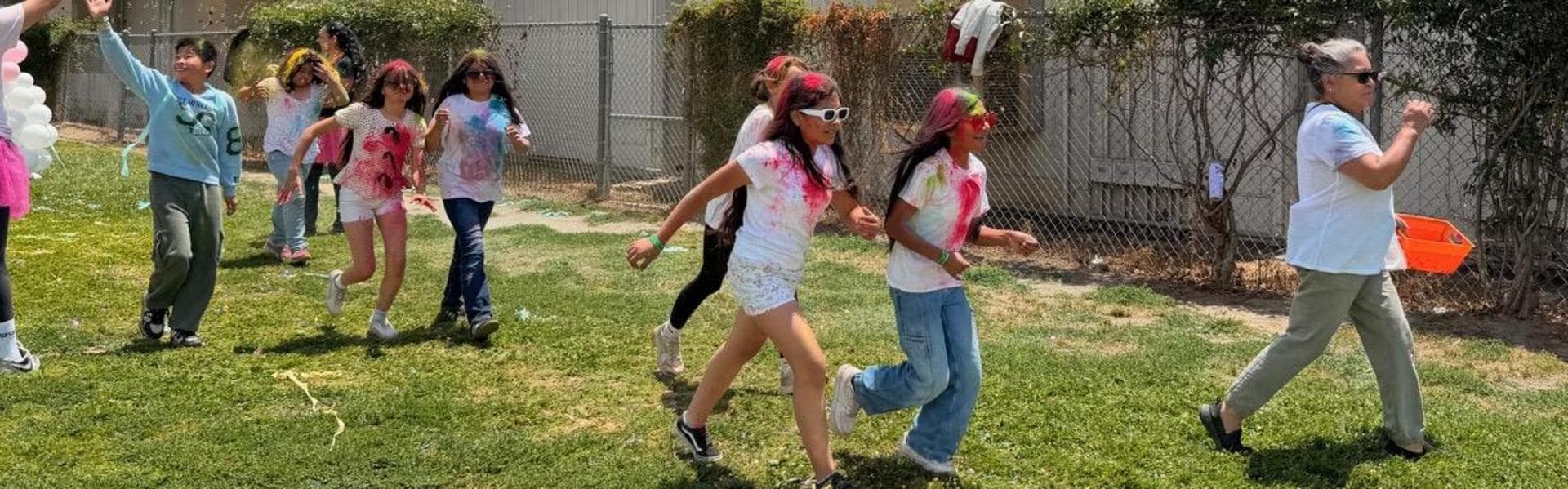Children walk joyfully in a line with colorful powder and balloons in an outdoor setting.