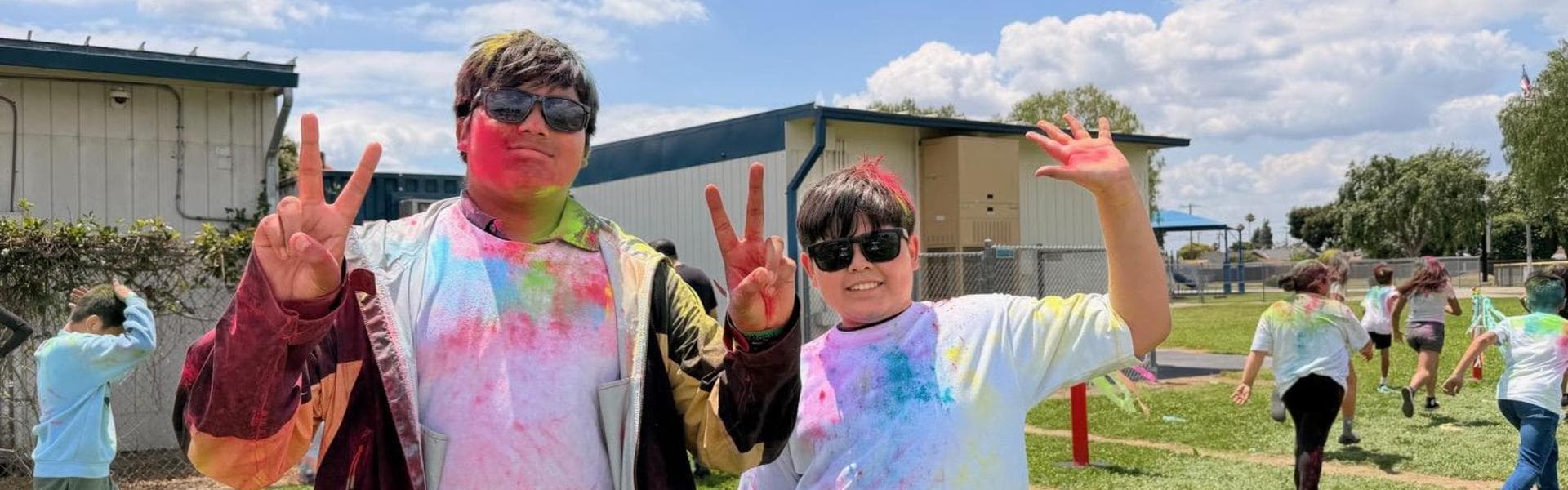 Two boys show peace signs, covered in colorful powder, against a school backdrop.