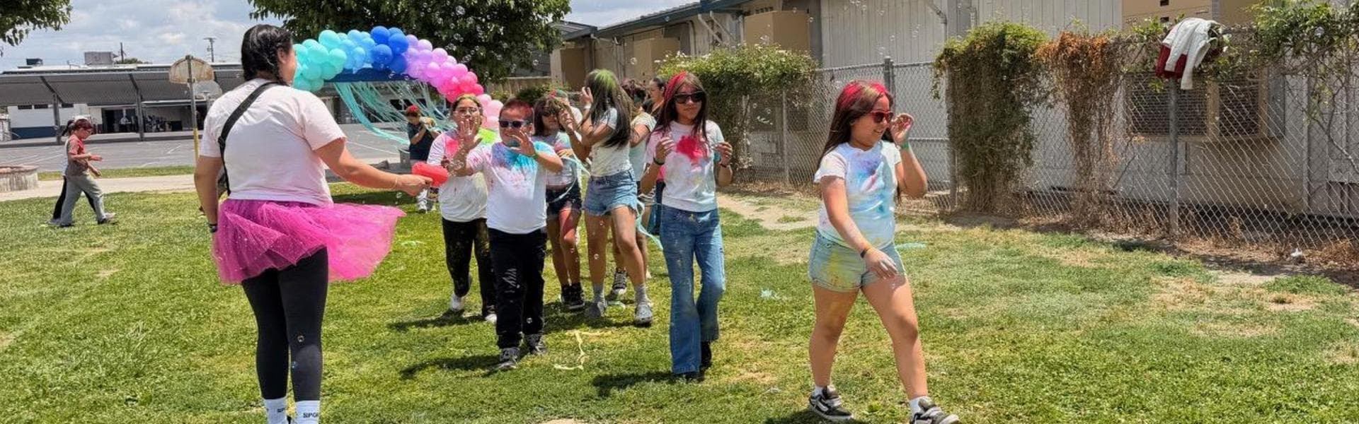 A group of kids in colorful clothes and sunglasses stroll on grass, with balloons nearby.