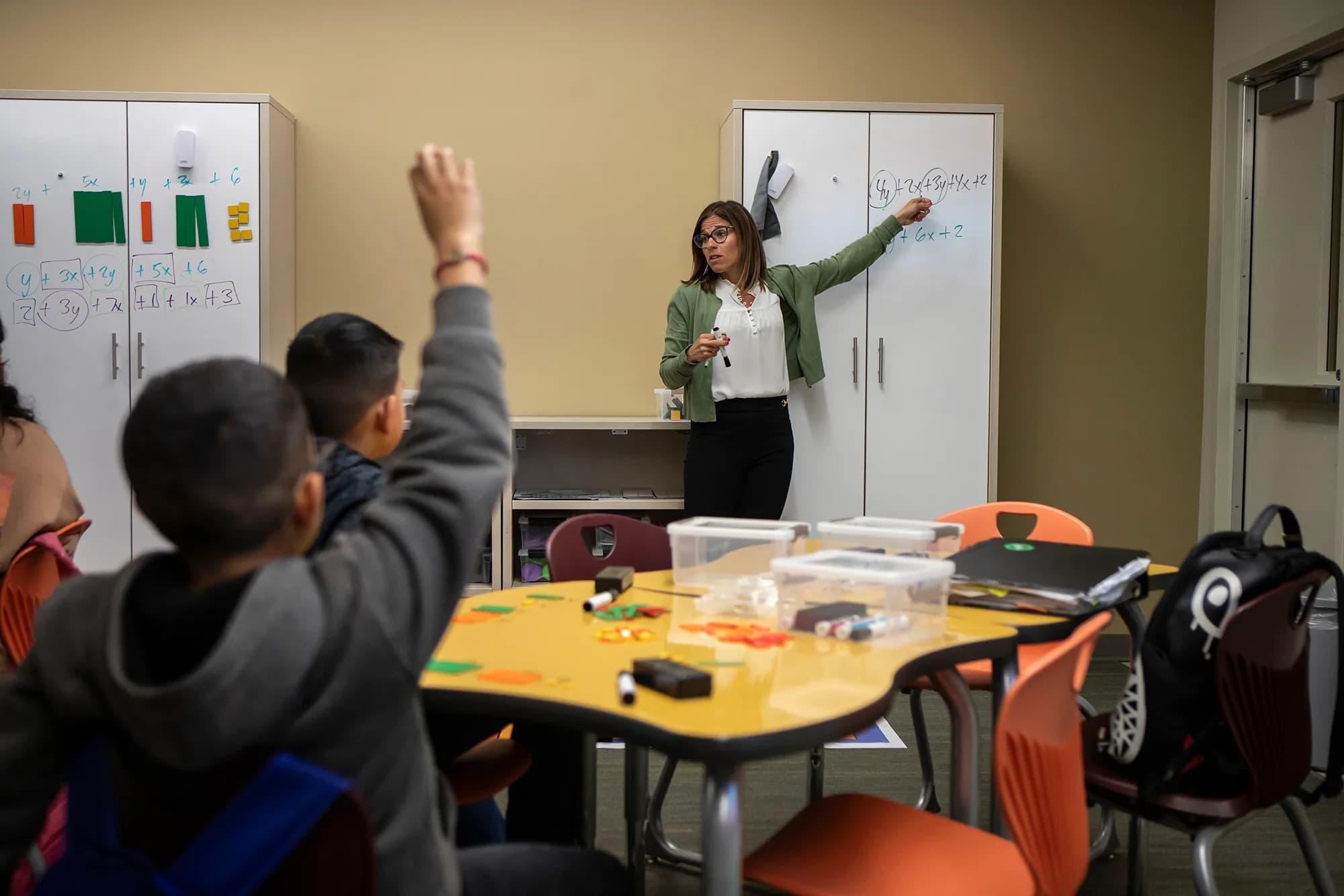 A classroom setting with a teacher standing at the whiteboard and students seated at desks, some with their hands raised.