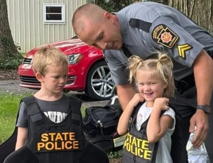 A uniformed police officer smiling and talking with two young children wearing black vests labeled 'State Police.' One child, a girl with blonde hair in pigtails, is smiling and has her fists raised in a playful pose. The boy is looking at the girl with a neutral expression. They are outdoors, with a red car and a building in the background.