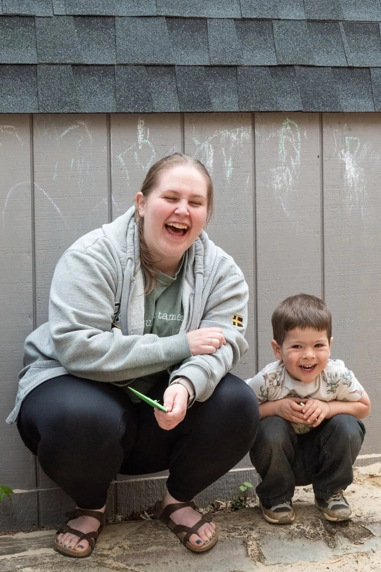 A woman and a young boy crouching outdoors in front of a gray wall, laughing and smiling with their eyes closed. The woman is wearing a gray hoodie, black pants, and sandals, holding a green object. The boy is wearing a beige shirt with a pattern and dark pants, holding a small object, and has short brown hair.