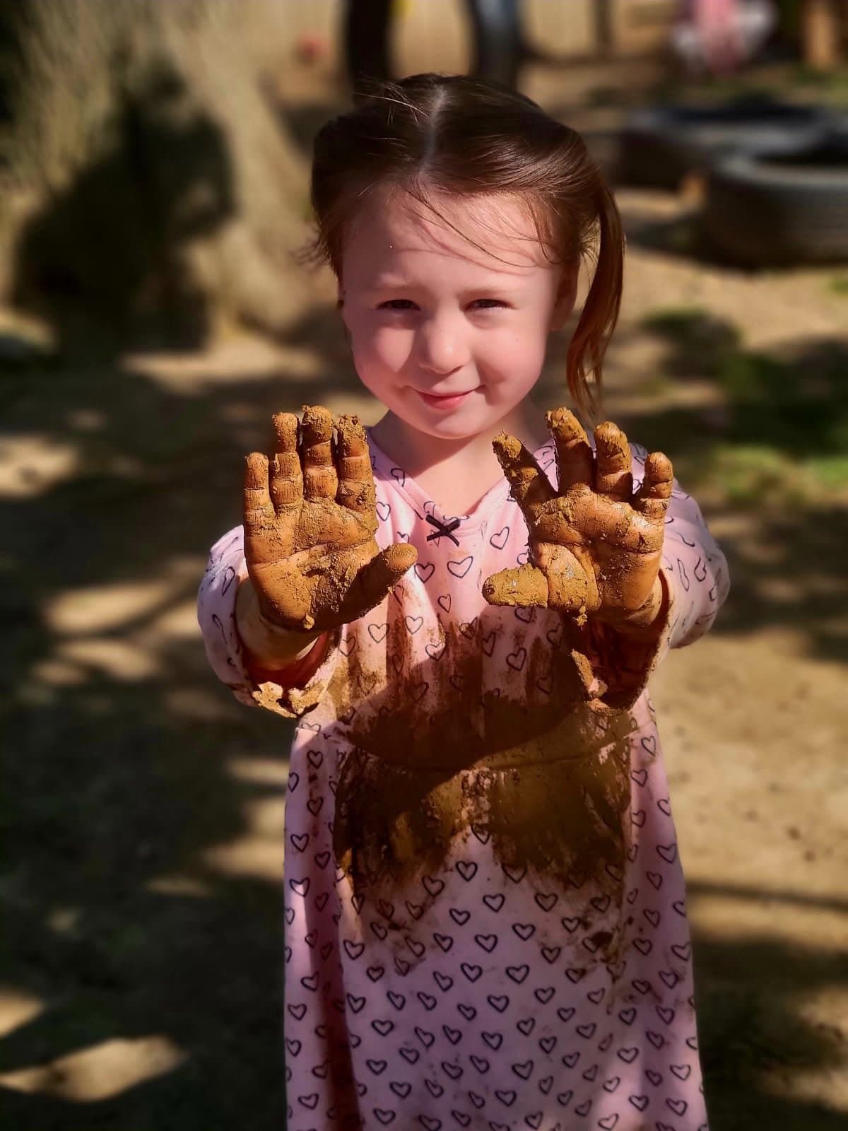 A young girl with brown hair in pigtails, wearing a pink dress with heart patterns, is showing her muddy hands and smiling outdoors.