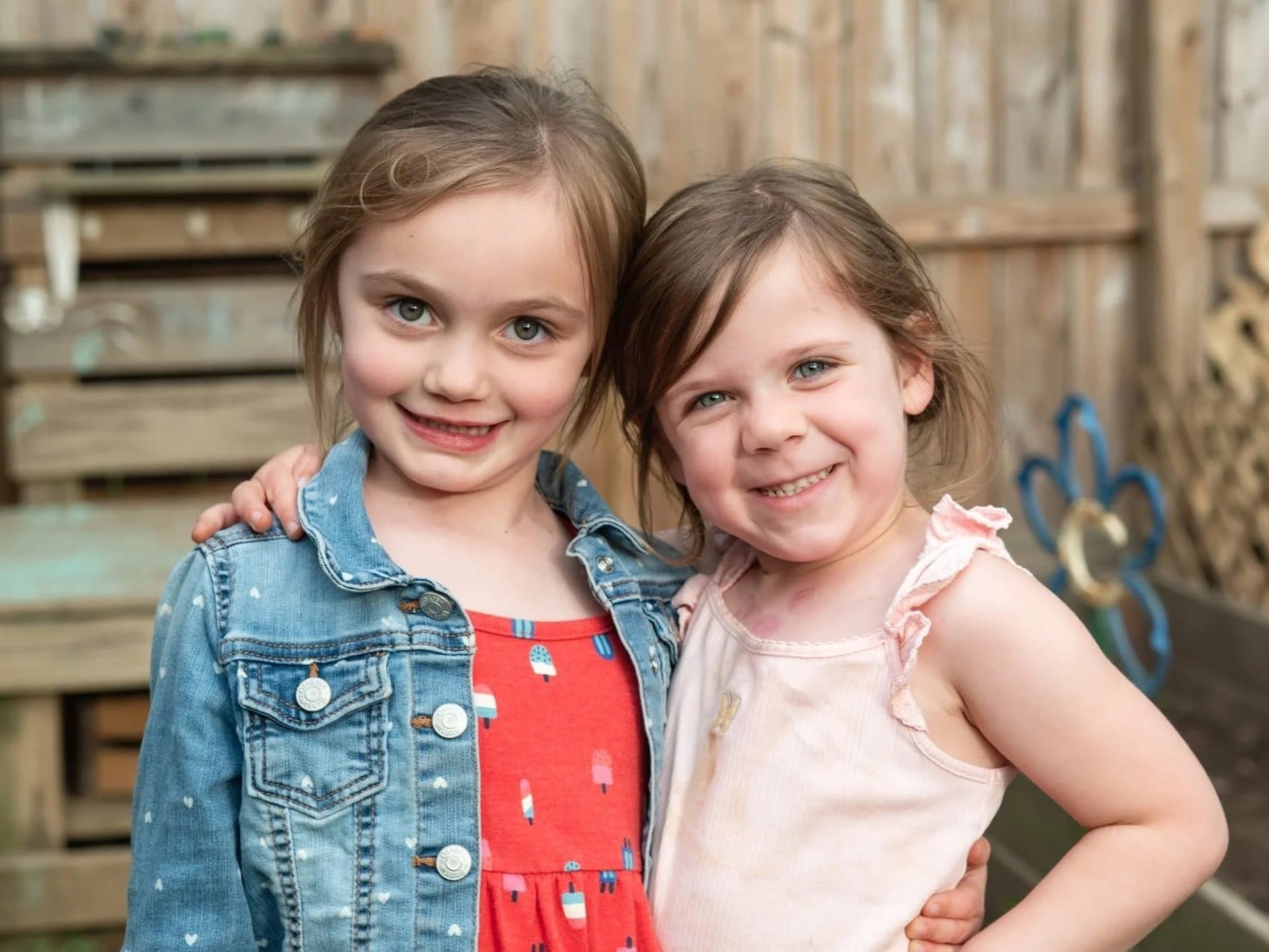 Two young girls standing close together outside, smiling at the camera, with a wooden fence and some children's play equipment in the background.