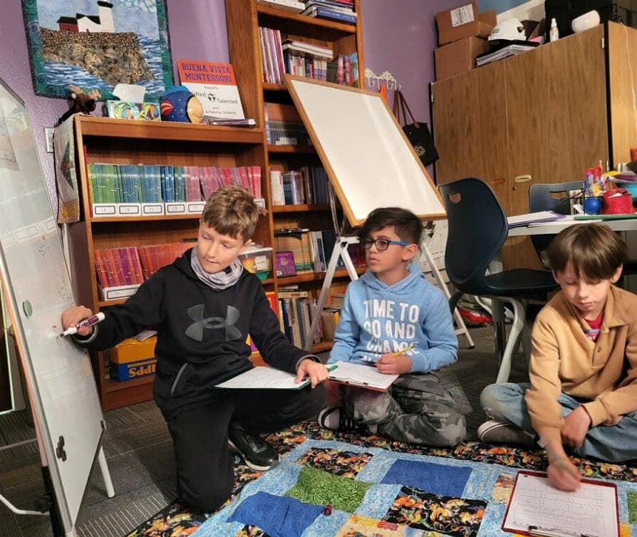 A male student teaches a concept on the whiteboard to two other male students.
