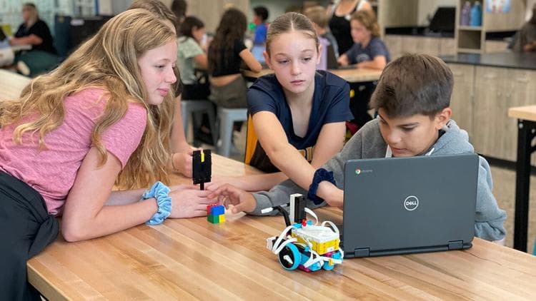 A group of young students, likely in a classroom setting, are gathered around a laptop computer, engaged in what appears to be a collaborative learning activity or project.