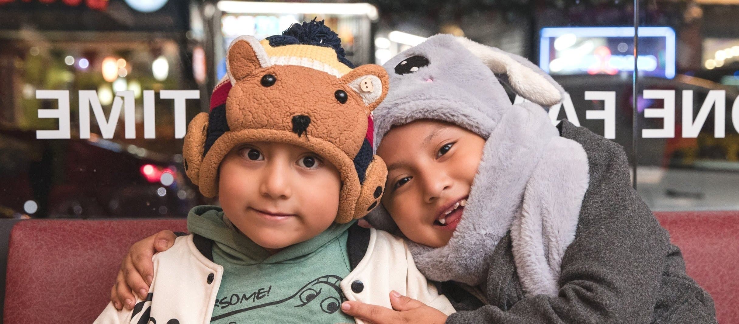 Two young children in animal hats sitting together indoors at night. One wears a bear hat and the other wears a gray bunny hat. They are smiling, with the bunny-hat child showing a tongue peek, and are close together.