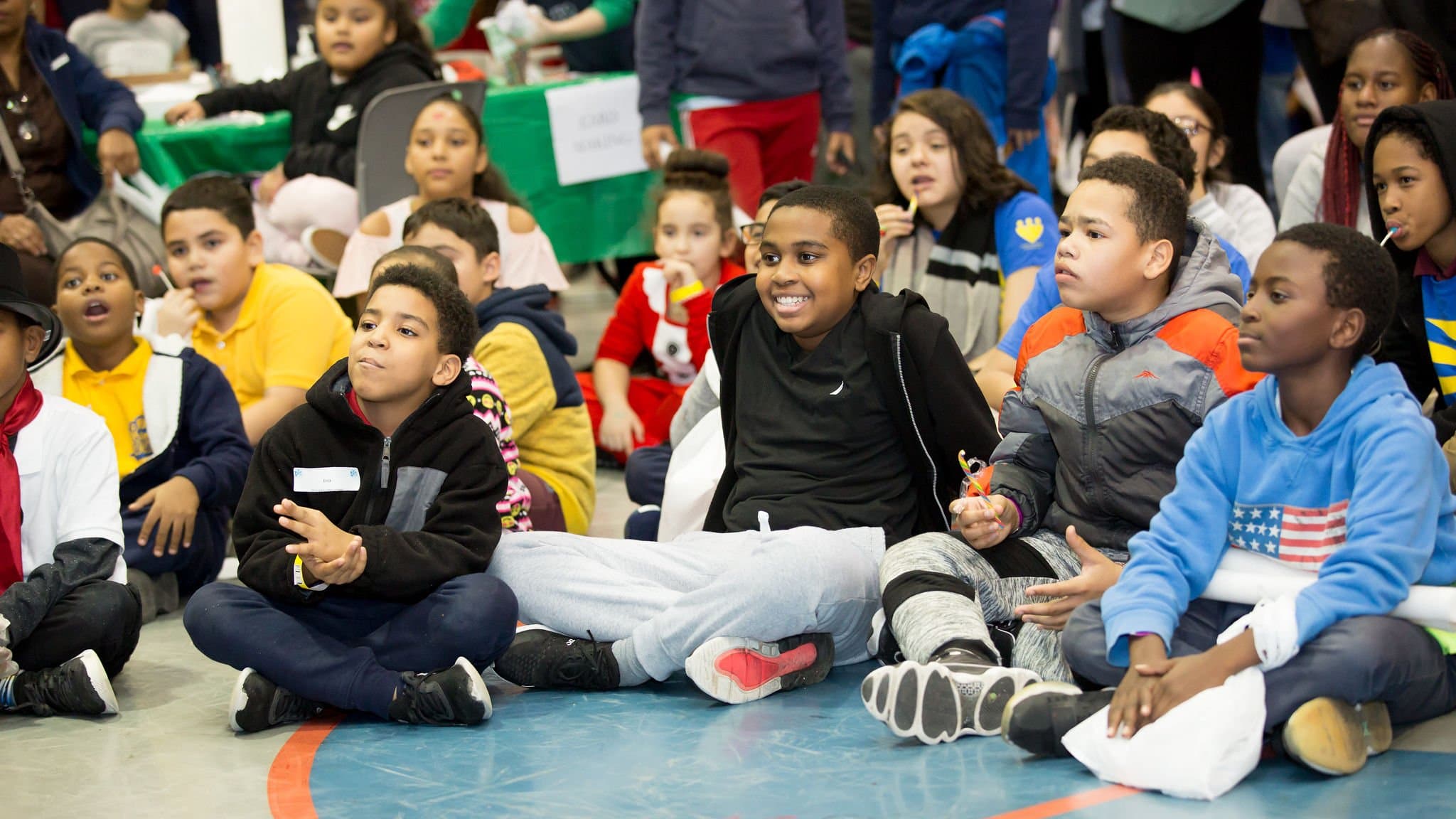 Children sitting on the floor at an indoor event, some smiling, some focused, with tables and decorations in the background.