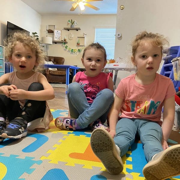 Three young girls sitting on a colorful play mat indoors.