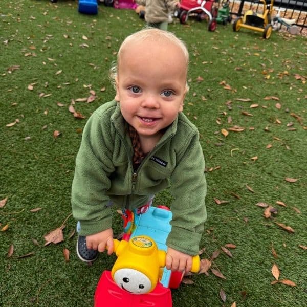 A smiling toddler riding a colorful toy bike outdoors on grass.