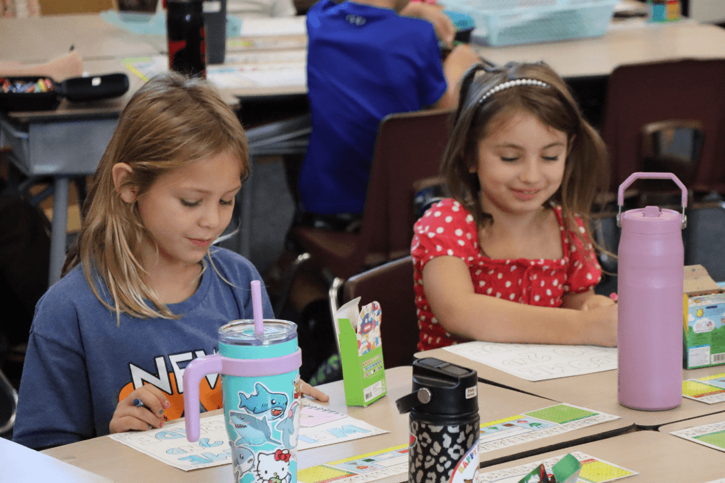 Two young girls work on drawings at their desks in a classroom.