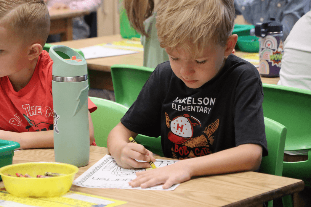 A young student concentrates on coloring at a desk in a classroom.