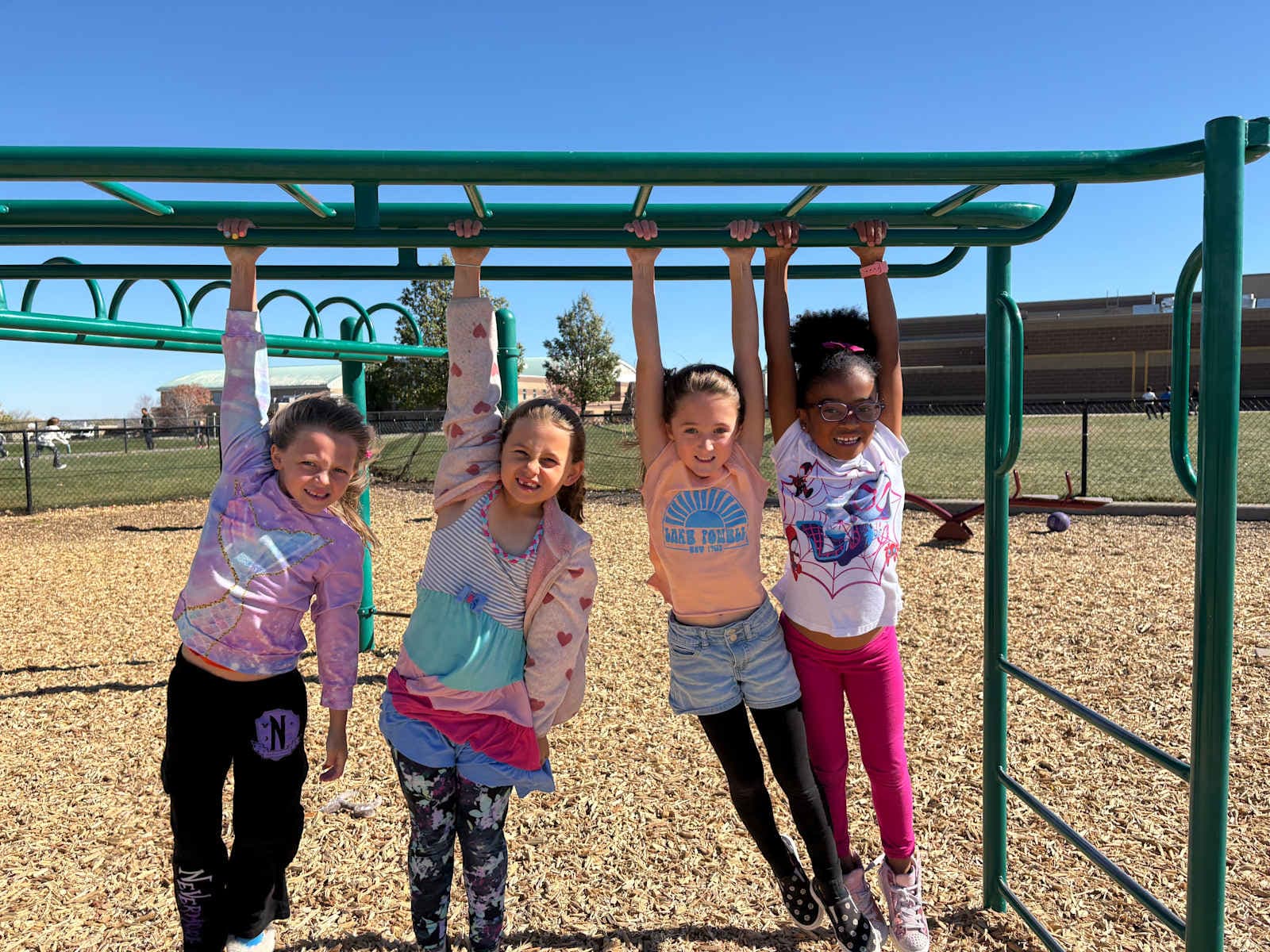 A group of young girls, dressed in colorful clothing, are hanging from the bars of a playground structure against a backdrop of a clear blue sky.