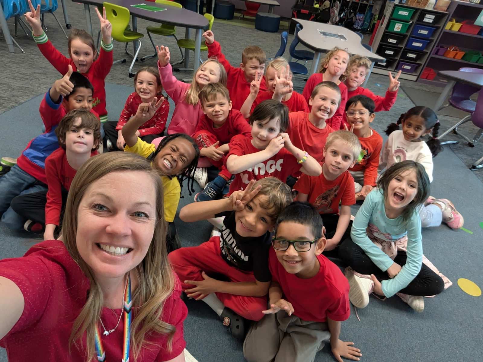 A group of young children wearing red shirts are gathered together in a classroom setting, with various educational materials and furniture visible in the background.