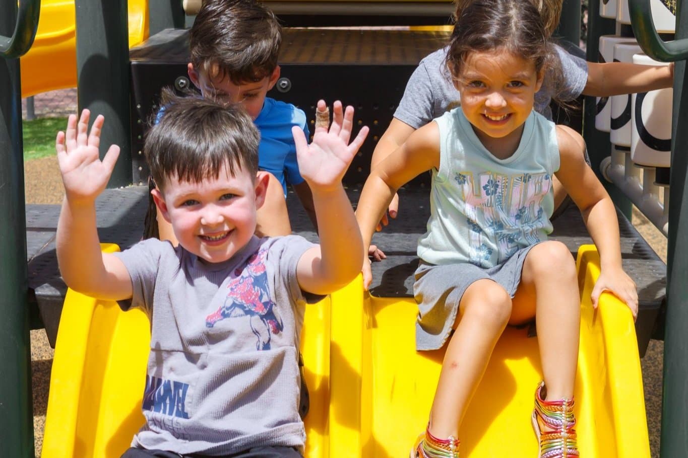 Four children playing on a yellow slide at a playground, smiling and waving at the camera.