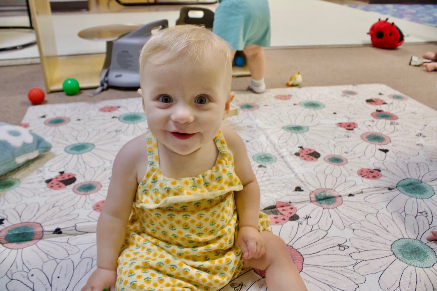 A smiling baby lying on a colorful play mat with a pink cartoon animal and various toys around, smiling and reaching forward.
