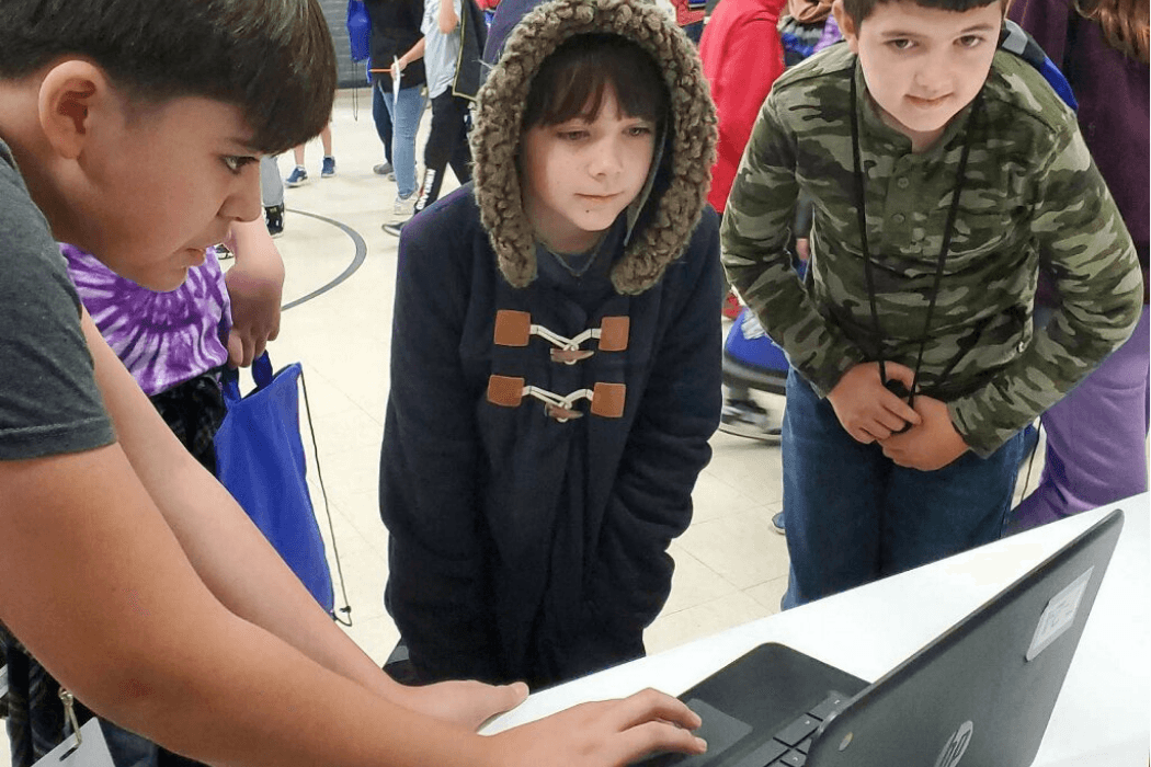 Three children look at a laptop computer, possibly working on a project.