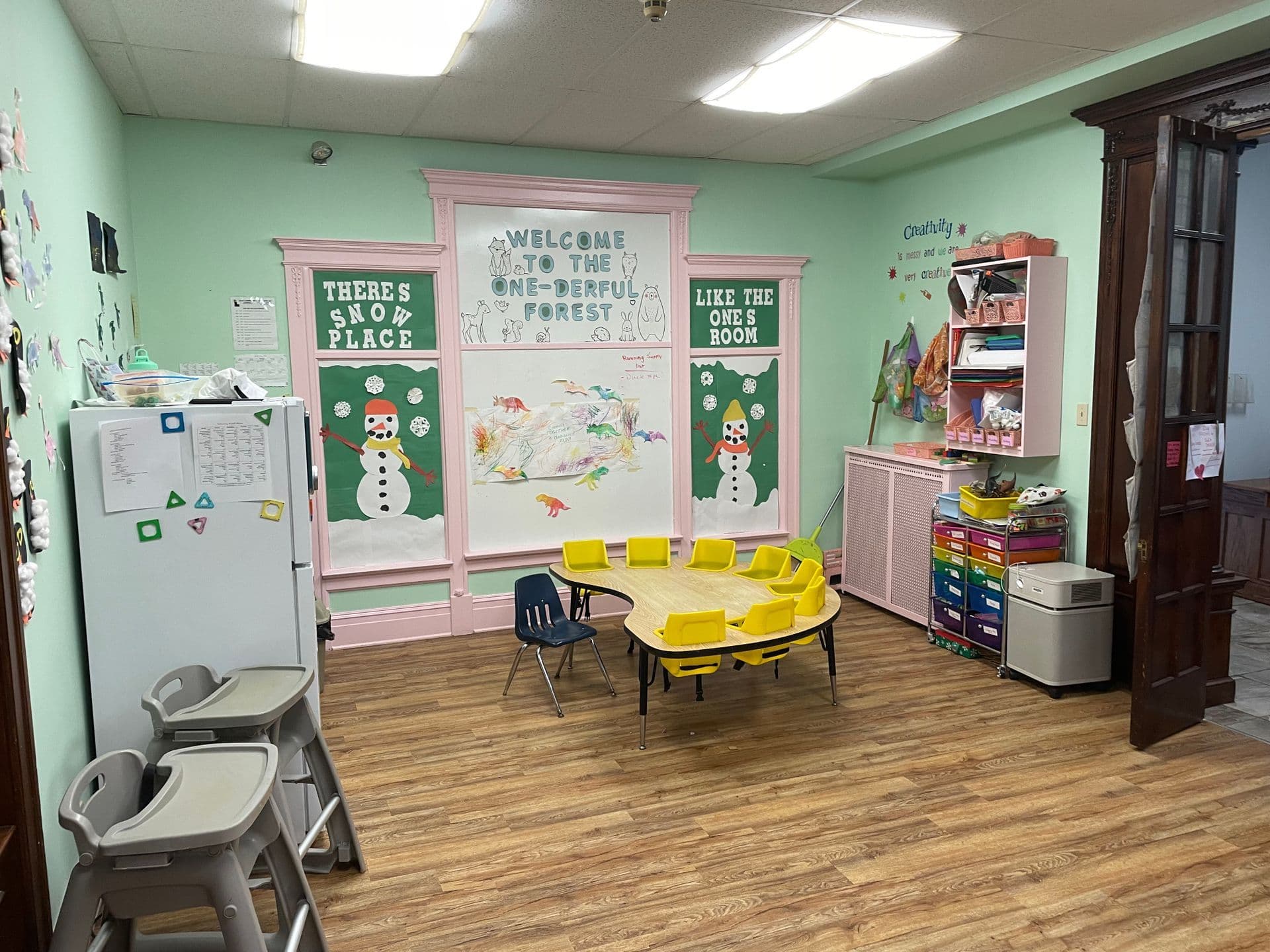 Empty classroom with mint walls, wood floor, child-sized tables and chairs, and colorful bulletin boards and shelves
