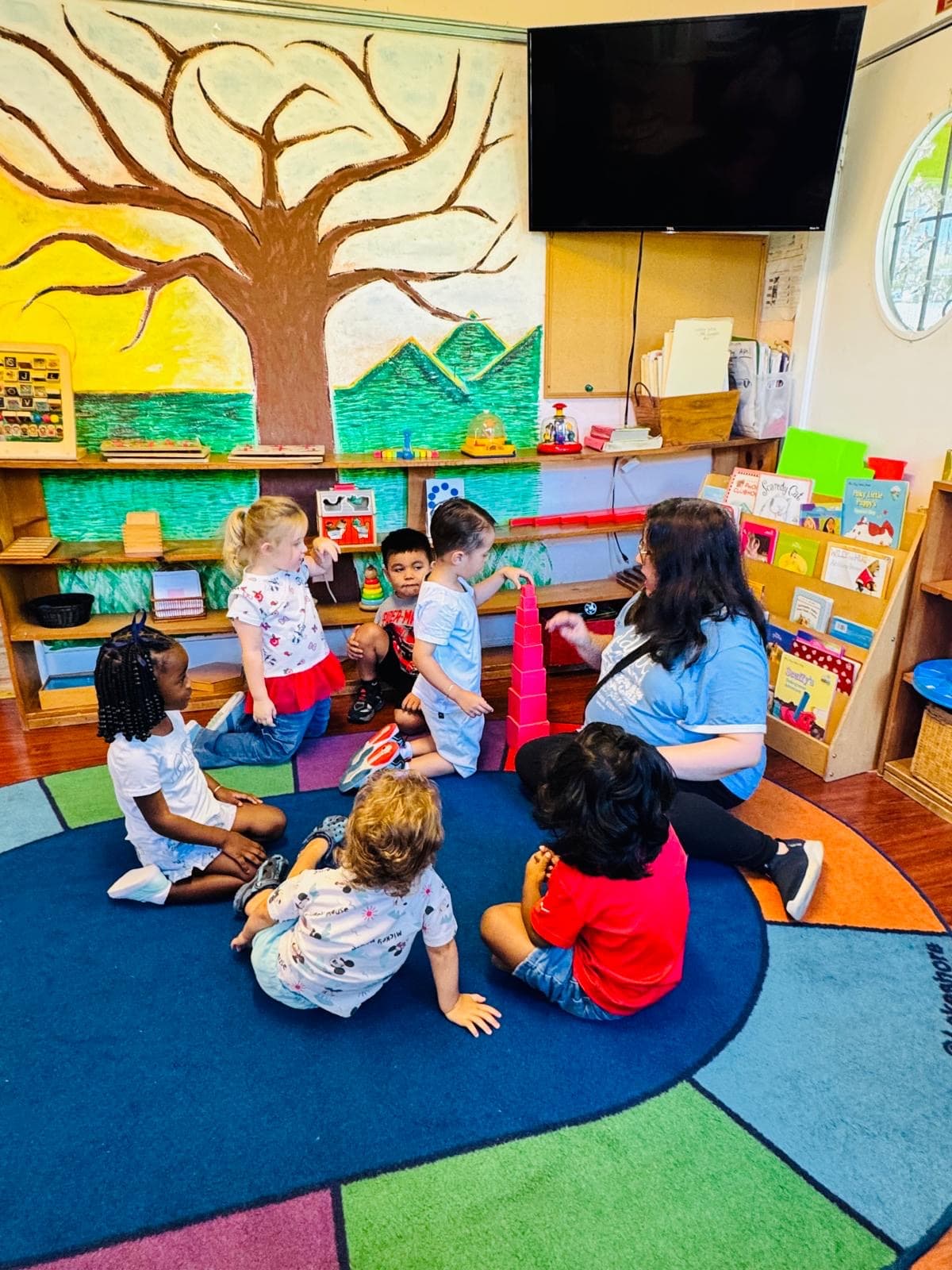 A teacher presents the pink tower to a mixed-age circle beneath the tree mural