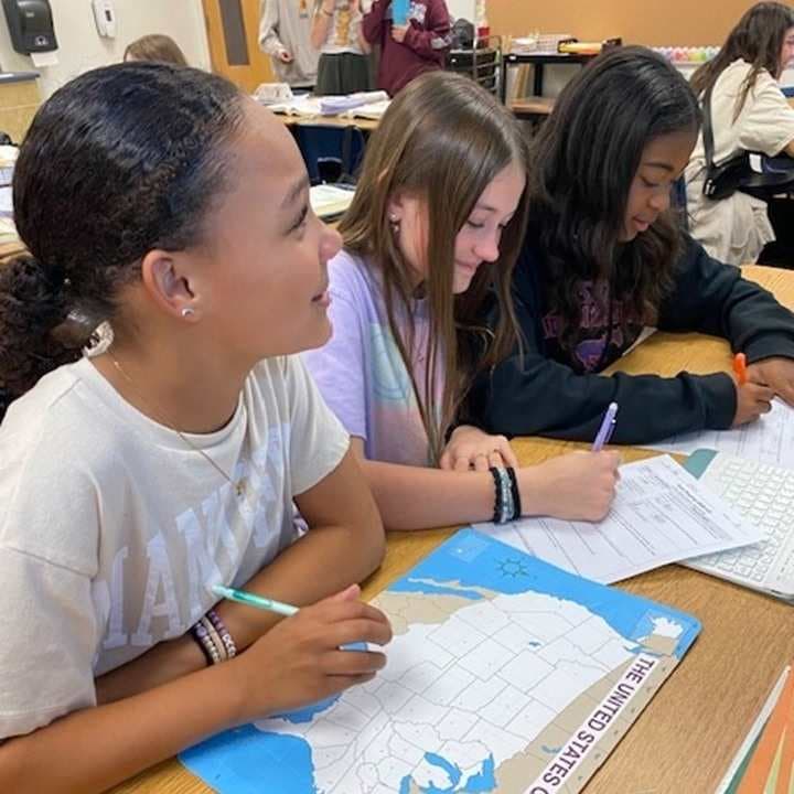The image shows several students working together at a desk, engaged in what appears to be a collaborative learning activity. The classroom environment in the background suggests an educational setting.