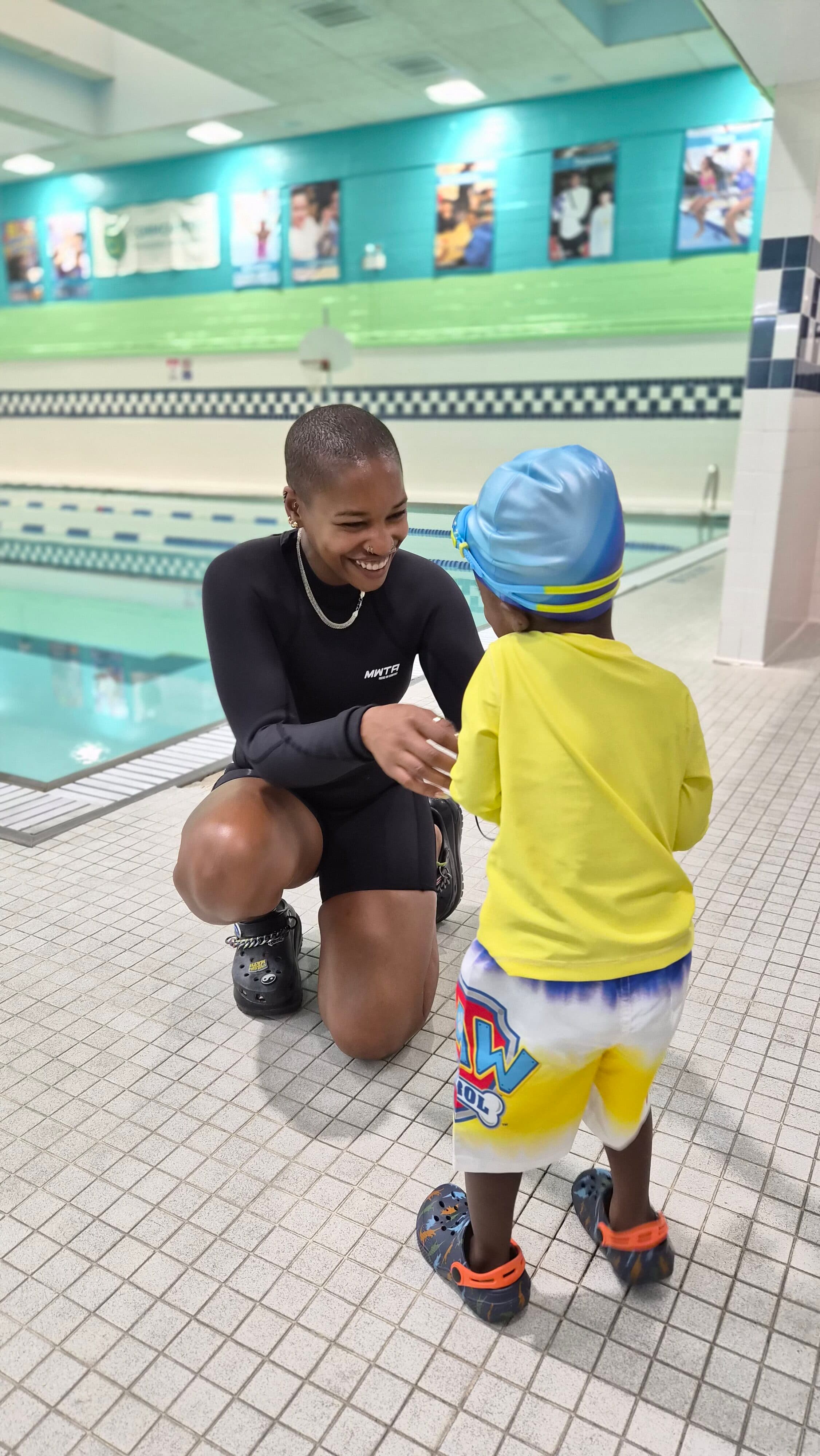 An adult in a black swimsuit kneels and smiles at a young child in swim gear on the deck of an indoor swimming pool, capturing a heartwarming moment during Bronx Aquatics swim classes.