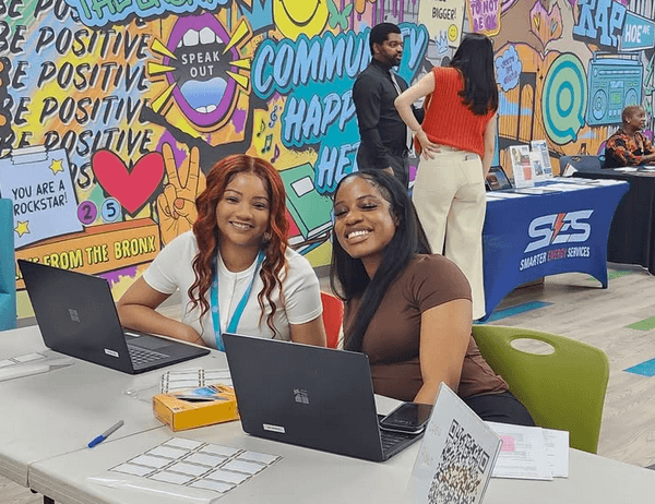 Two women sit at a table with laptops, smiling at the camera. Behind them is a colorful mural and a few people standing near informational booths.