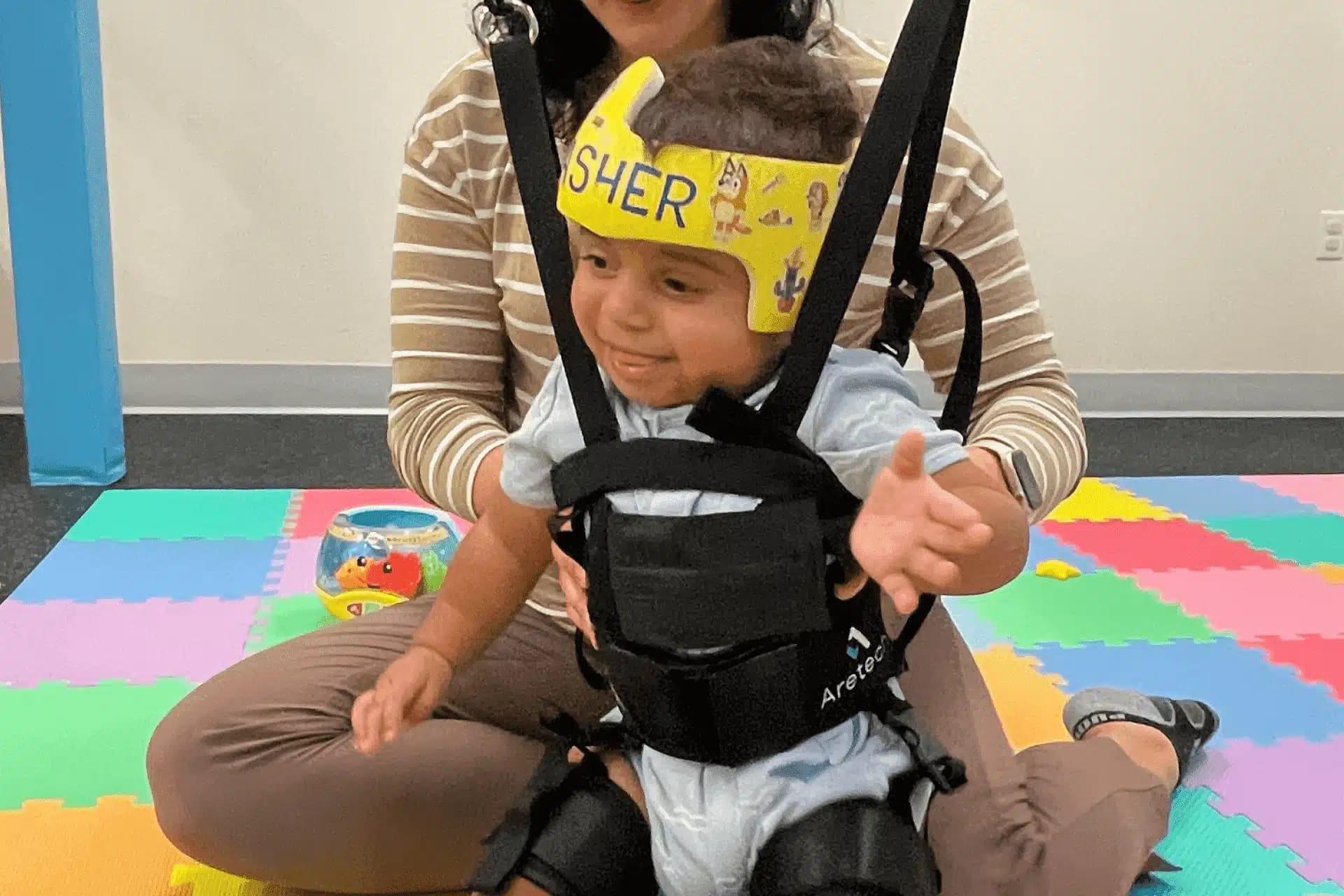 A young child wearing a yellow headband and harness sits on a woman's lap on colorful foam mats in a playroom.