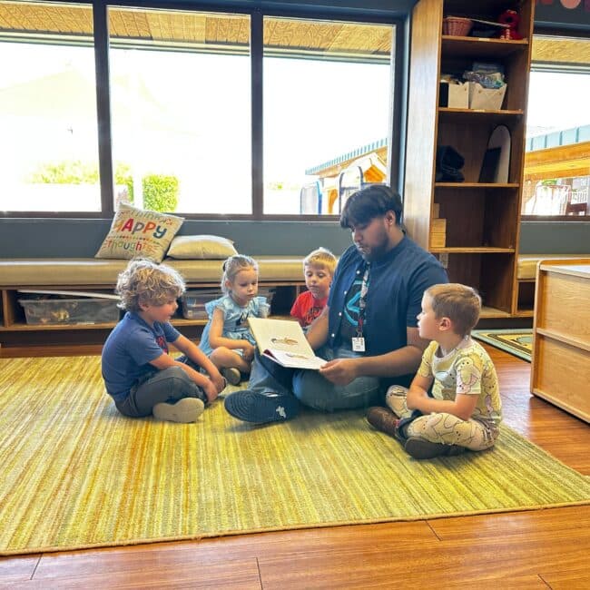 An adult reads a book to four young children sitting in a circle on a rug in a classroom with large windows.