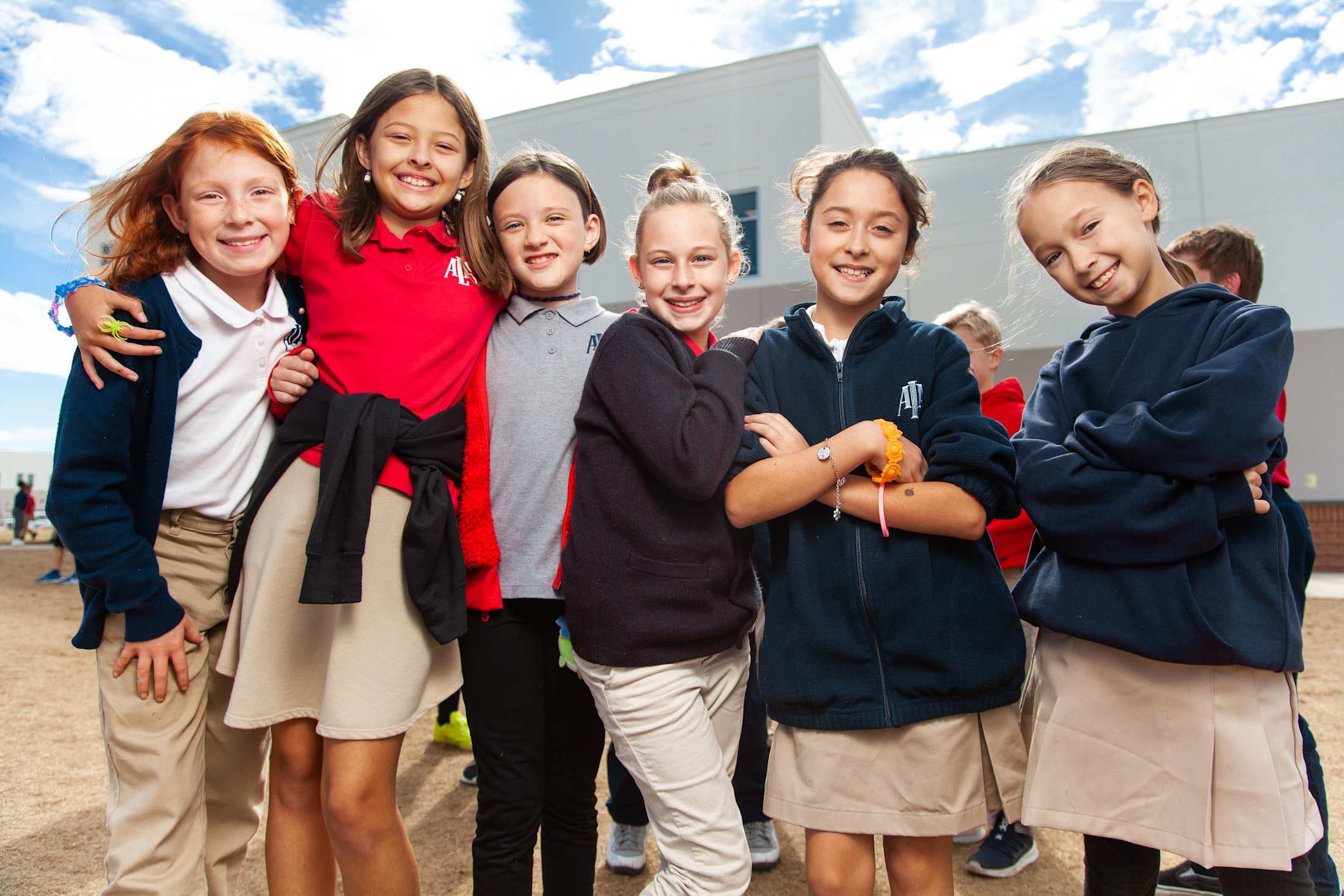 A group of smiling young students, dressed in school uniforms, stand together in front of a modern building with a cloudy sky in the background.
