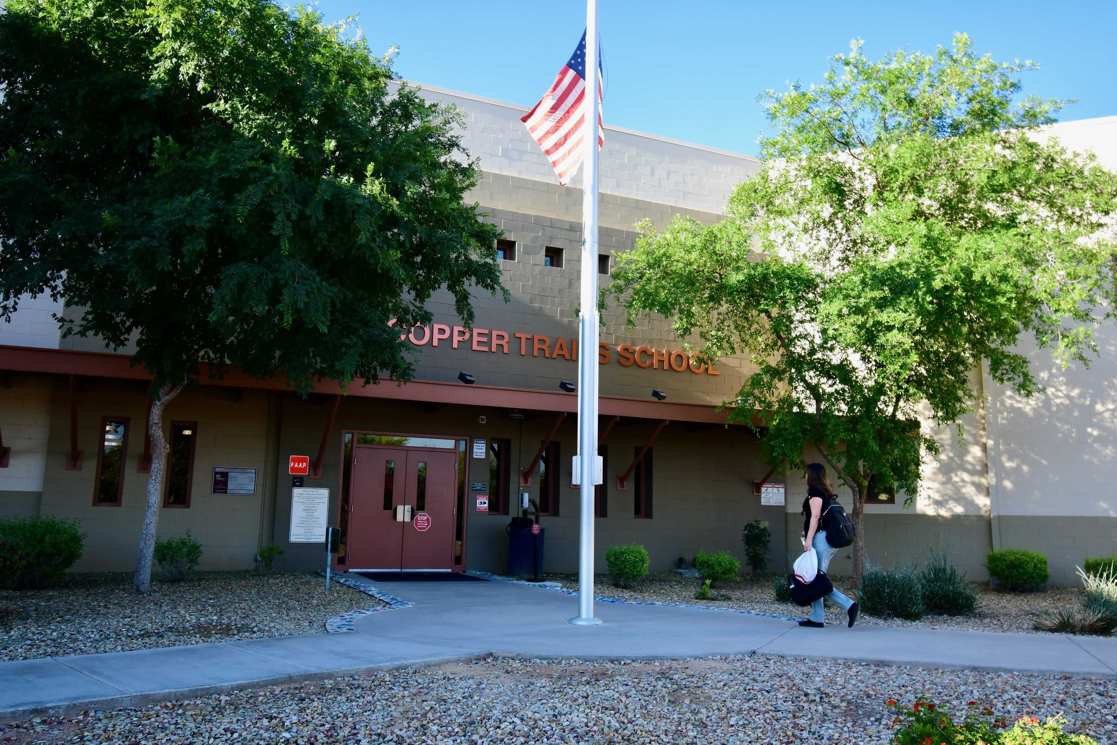 Photo of the front of Copper Trail School with trees, a flagpole and someone walking on the sidewalk
