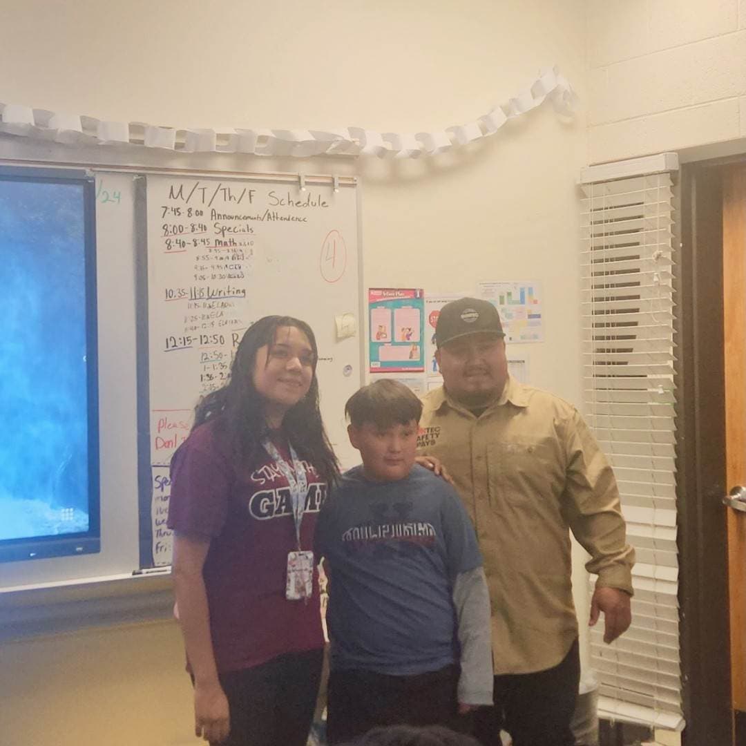 The image shows a woman and a young boy standing together in what appears to be a classroom or educational setting, with a whiteboard and other classroom materials visible in the background.