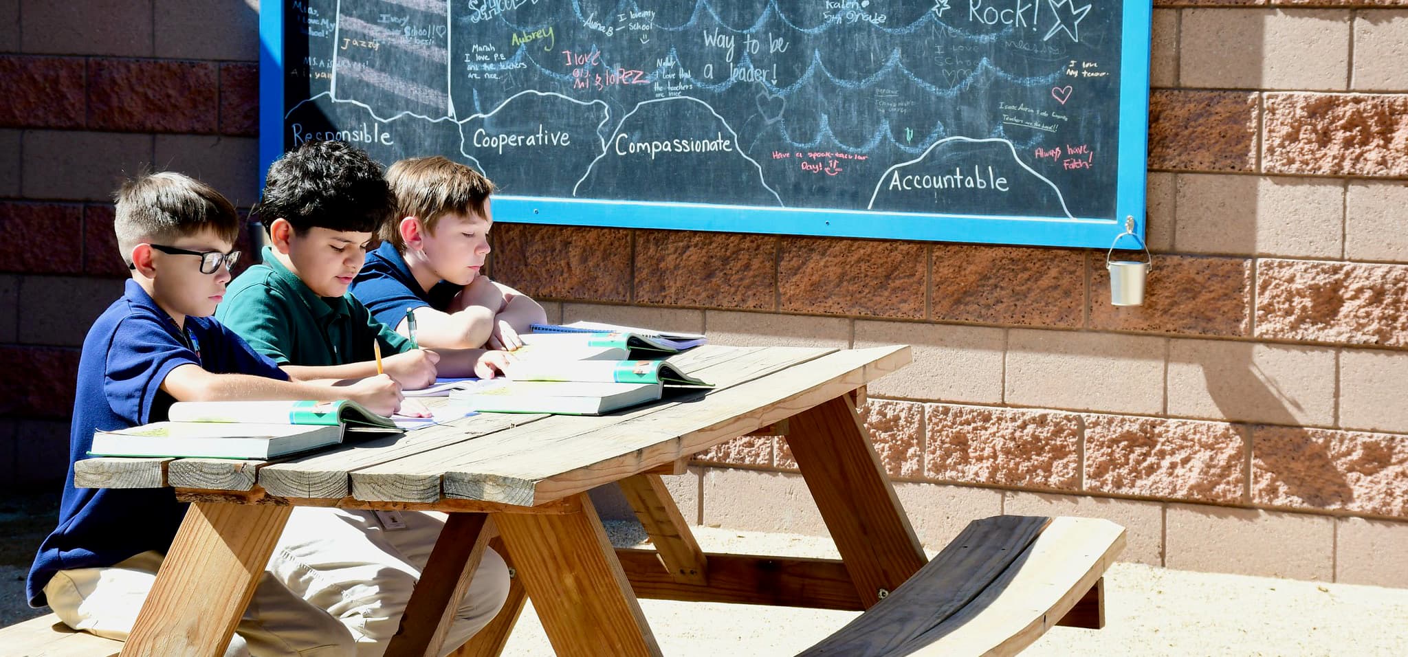 Three students sitting on a picnic table reading with a blackboard in the background with inspirational words